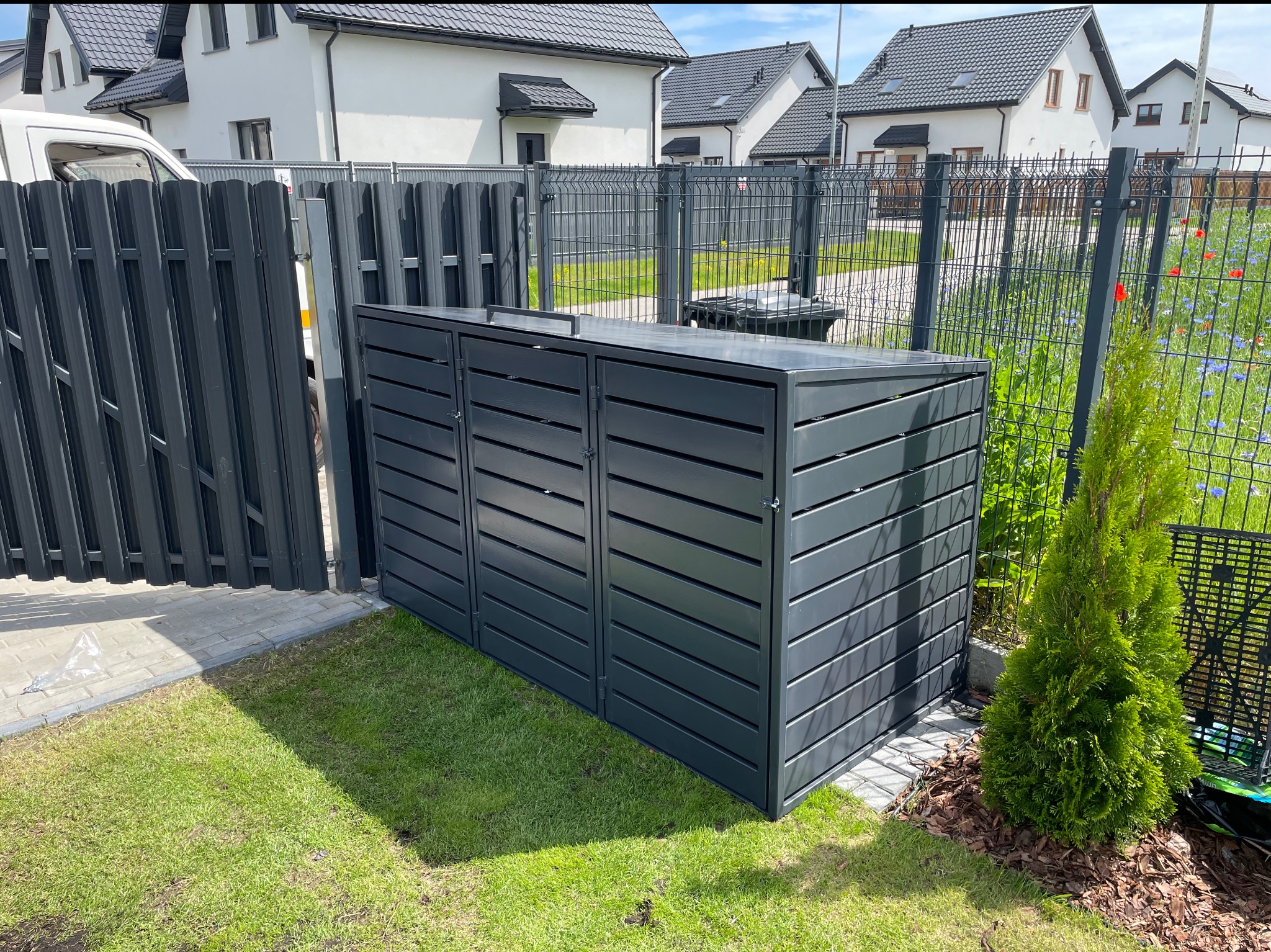 Nowoczesna zabudowa śmietnikowa z szarego metalu na trawniku, z widocznym ogrodzeniem panelowym i fragmentem ogrodzenia metalowego palisadowego.