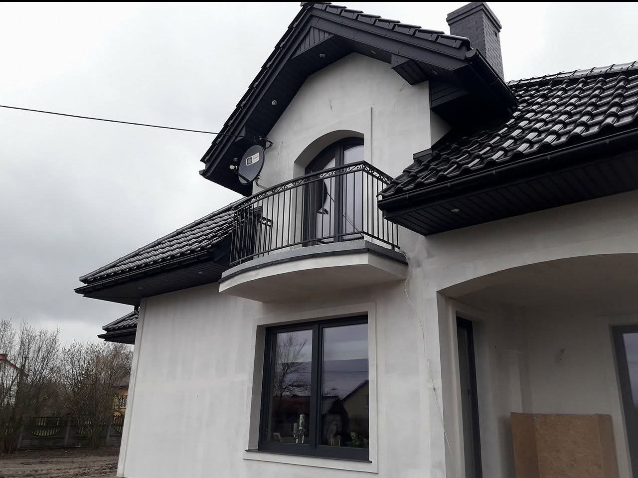 Elewacja domu z balkonem i czarną, metalową balustradą. Widoczna antena satelitarna i dach z czarną dachówką. Szara, pochmurna pogoda.