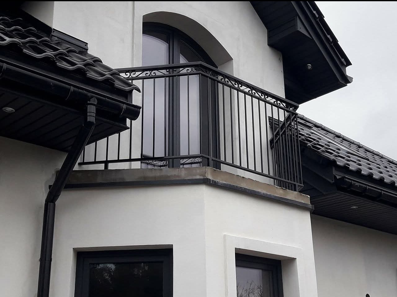 Czarna, metalowa balustrada balkonowa z dekoracyjnymi elementami na tle białej elewacji domu z czarną dachówką i oknami. Ujęcie z dołu, podkreślające detale.