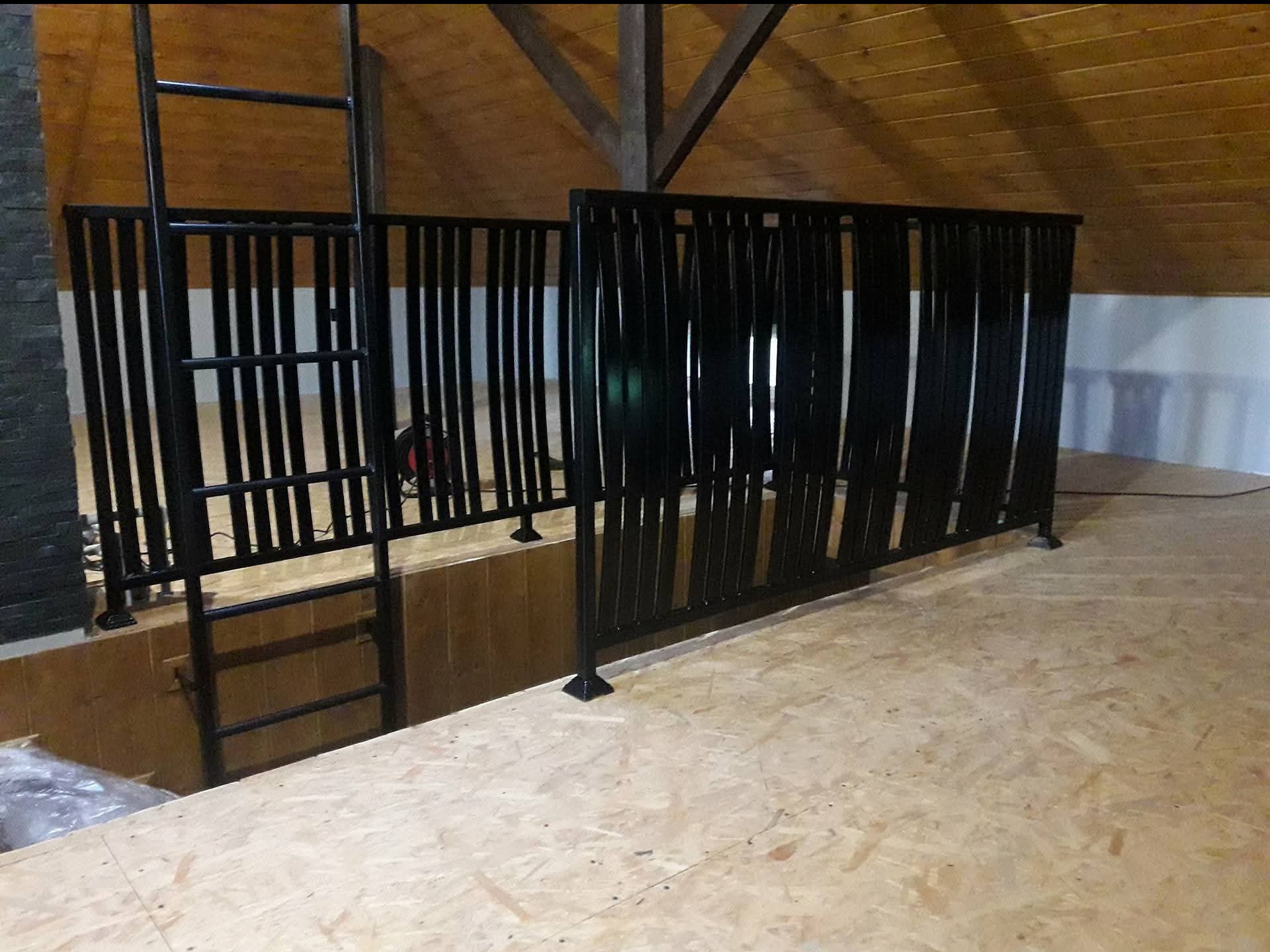 Czarna, metalowa balustrada z pionowymi szczeblami i drabinką, zamontowana na antresoli z drewnianym stropem. Widoczne spawy i mocowania.