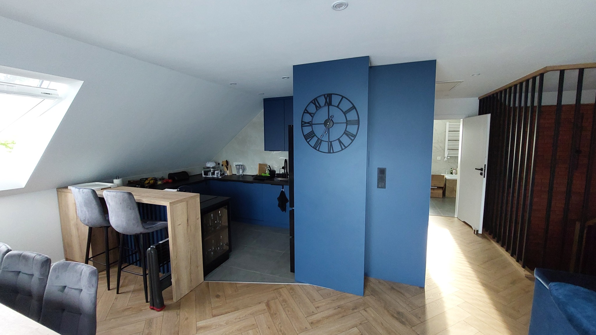 Niebieska kuchnia na poddaszu z zegarem na ścianie i drewnianym blatem barowym. Widoczny parkiet w jodełkę i fragment łazienki w tle.