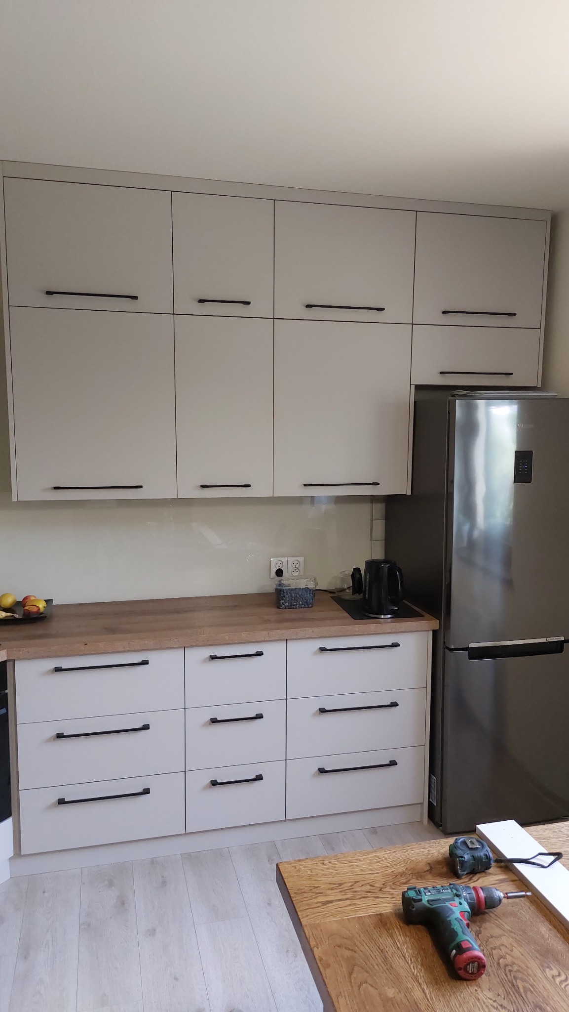 Jasna kuchnia z drewnianym blatem i czarnymi uchwytami, widoczna lodówka i narzędzia na stole, sugerujące montaż.