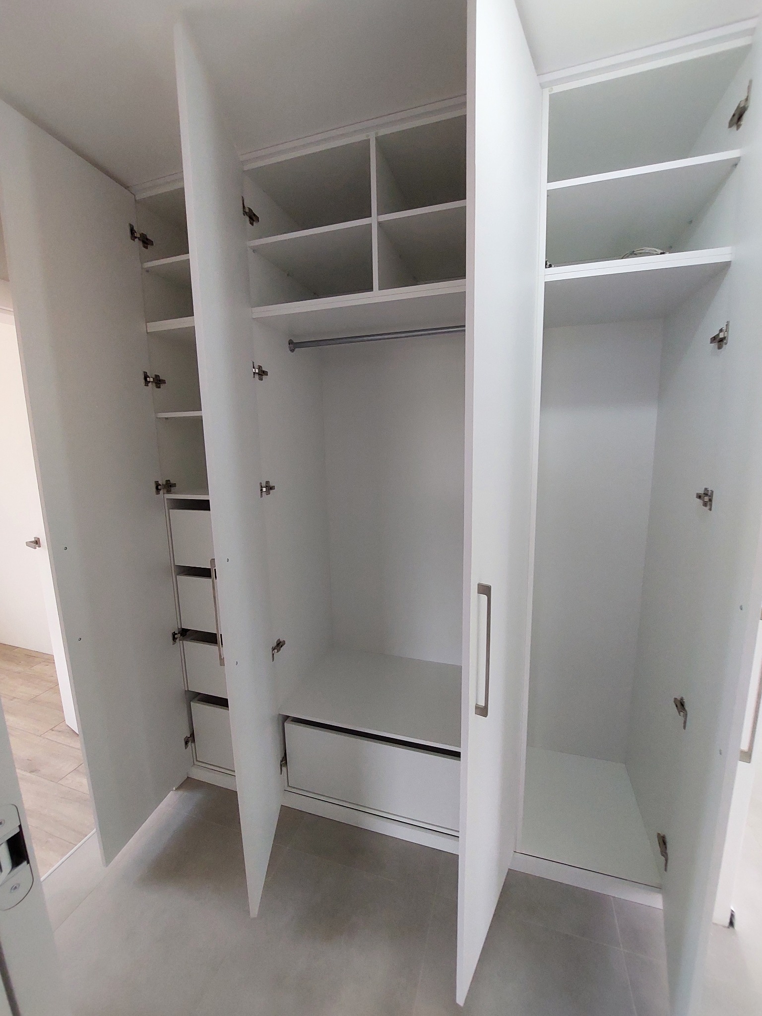 Biała szafa wnękowa z otwartymi drzwiami, widoczne półki, szuflady i drążek na ubrania. Minimalistyczny design, jasne wnętrze, szare płytki podłogowe.