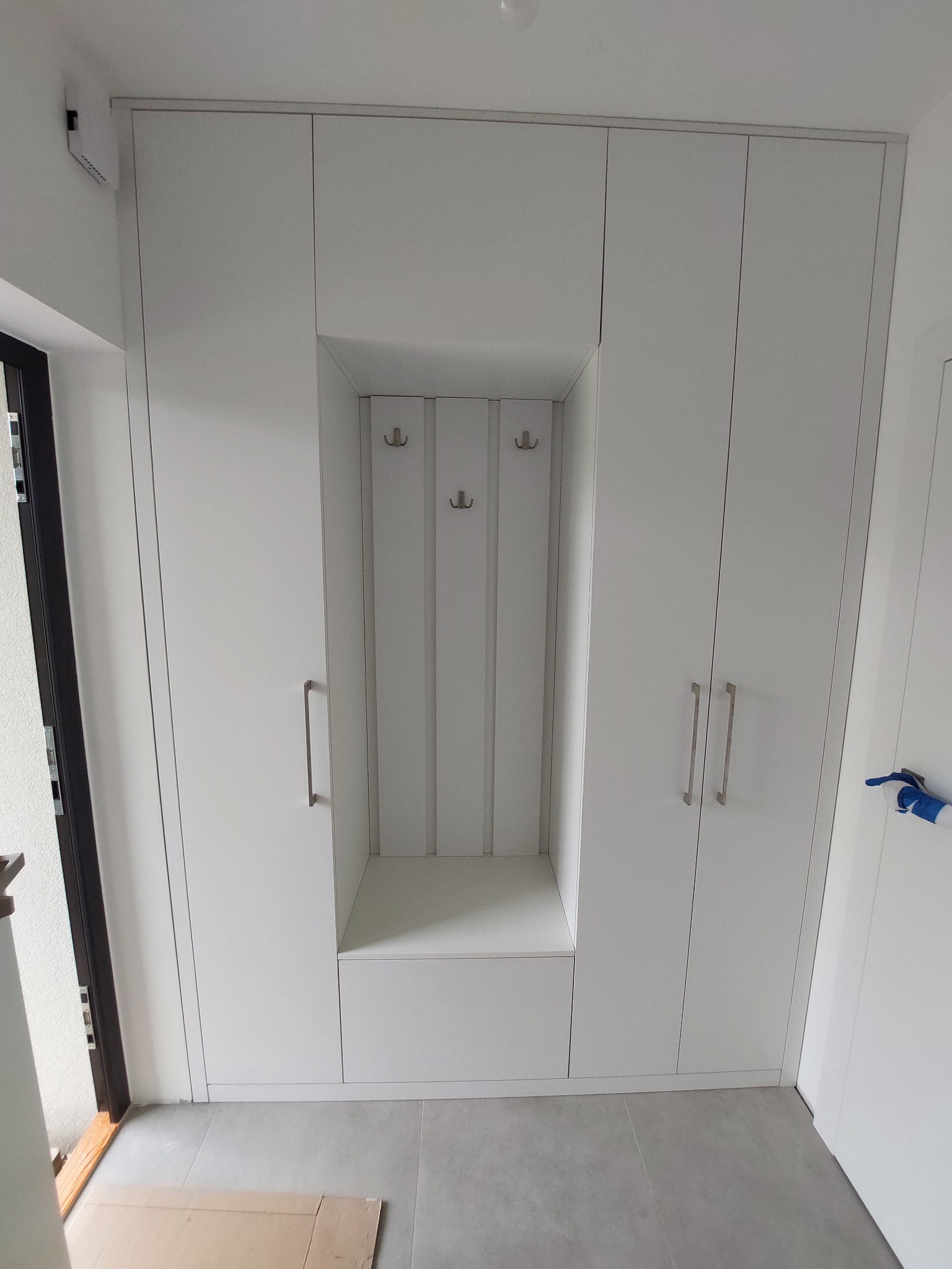 Biała szafa wnękowa z siedziskiem i wieszakami w przedpokoju. Minimalistyczny design, szare płytki podłogowe, nowoczesne wykończenie.