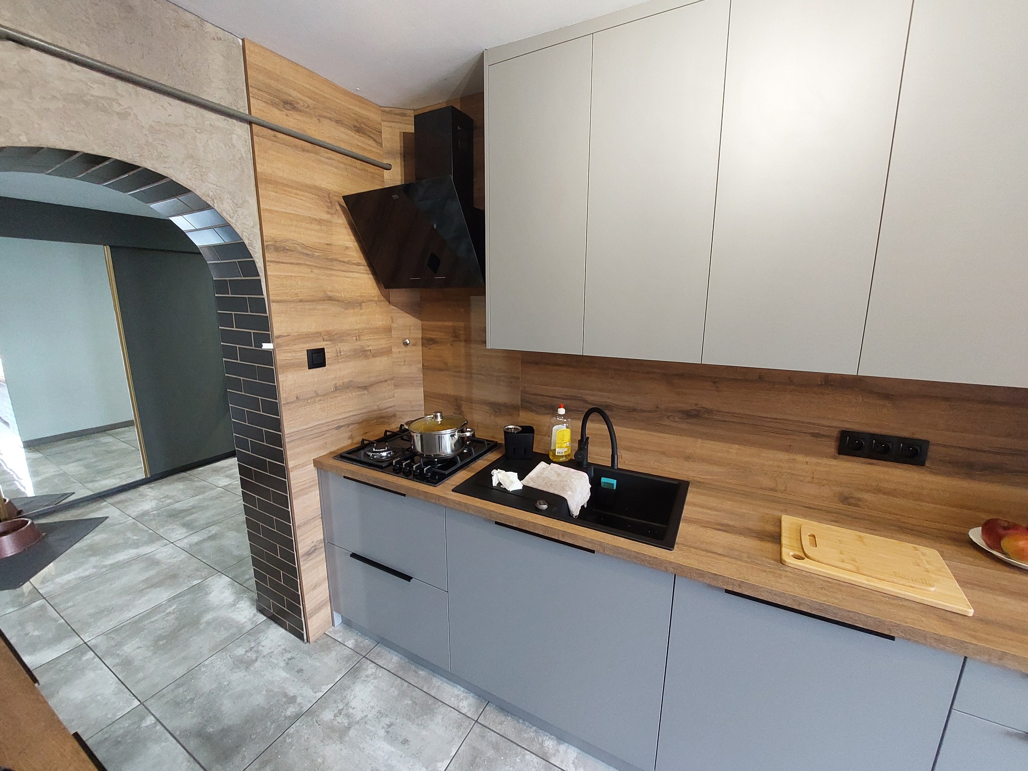 Szara, nowoczesna kuchnia z drewnianym blatem i czarnym okapem, widok na łukowe przejście do innego pomieszczenia, oświetlenie górne, płyta grzewcza z garnkiem.