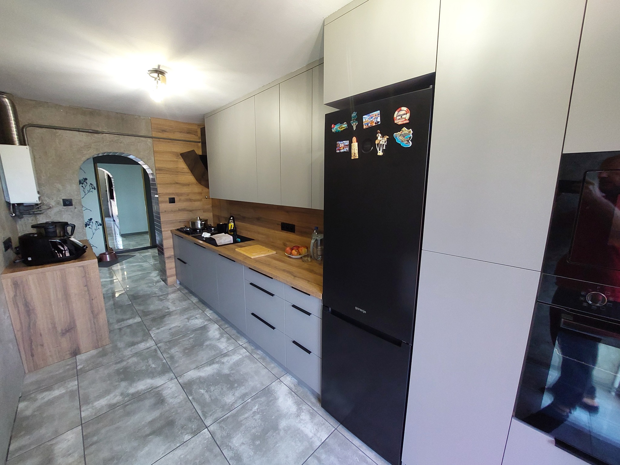 Szara kuchnia z drewnianym blatem i czarną lodówką, ozdobiona magnesami. Widoczny fragment piekarnika i podłoga z szarych płytek. W tle przejście do innego pomieszczenia.