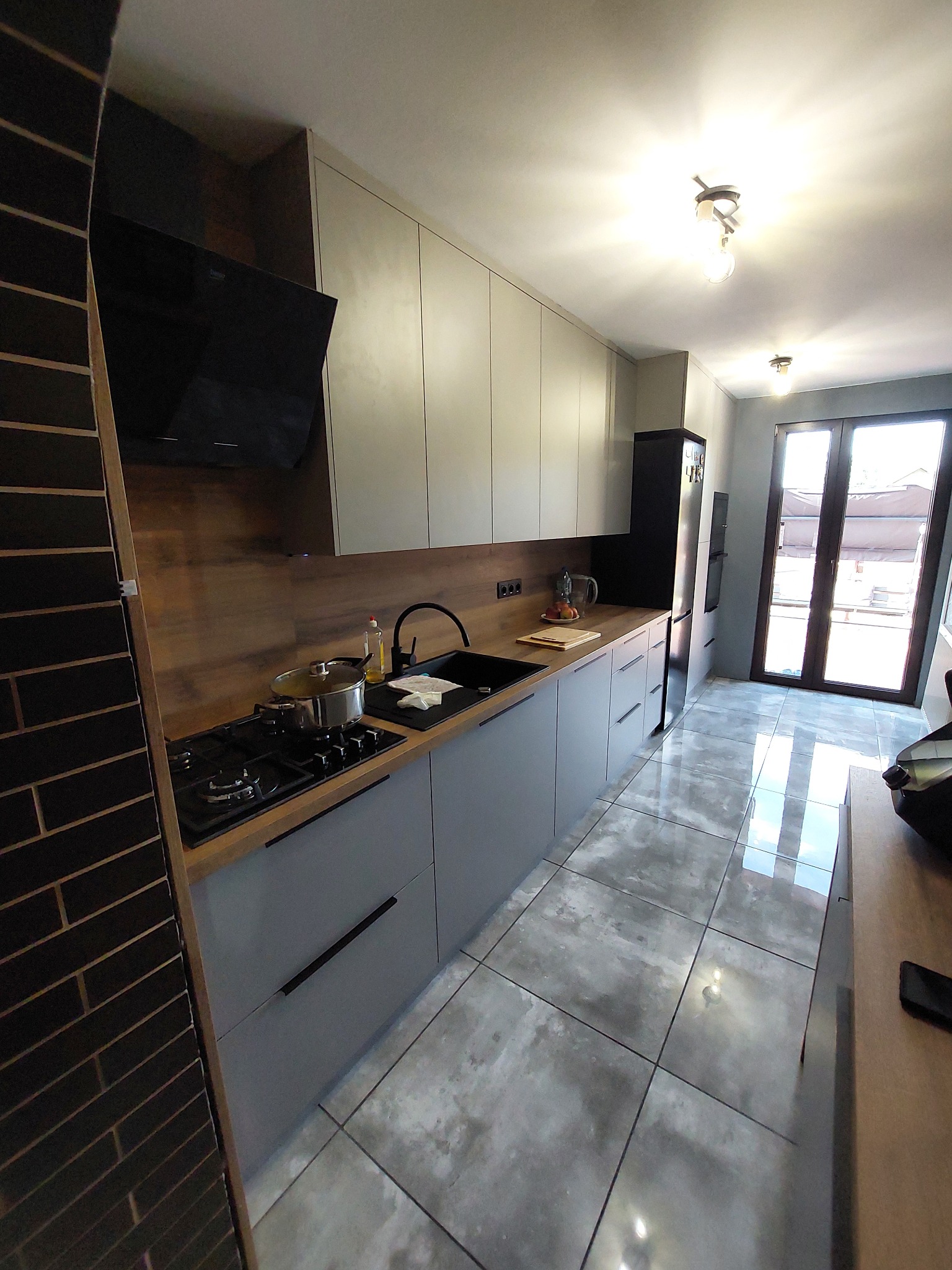 Nowoczesna kuchnia w odcieniach szarości z drewnianymi akcentami, czarnym sprzętem AGD i ceglaną ścianą. Widok na jasne drzwi balkonowe.