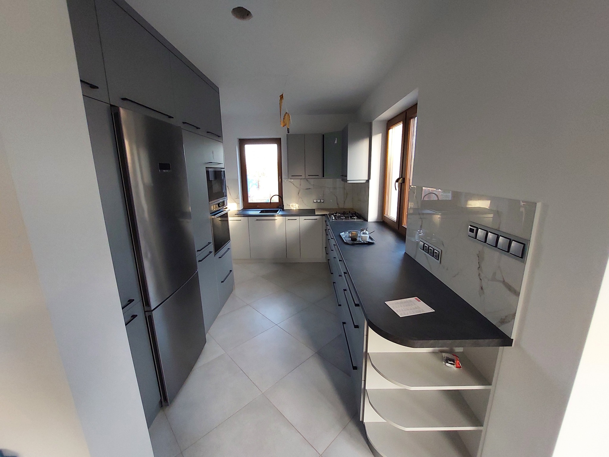 Nowoczesna kuchnia z szarymi szafkami, wyspą z półkami i marmurowym backsplashiem. Duża lodówka i okna z drewnianymi ramami dopełniają przestrzeń.