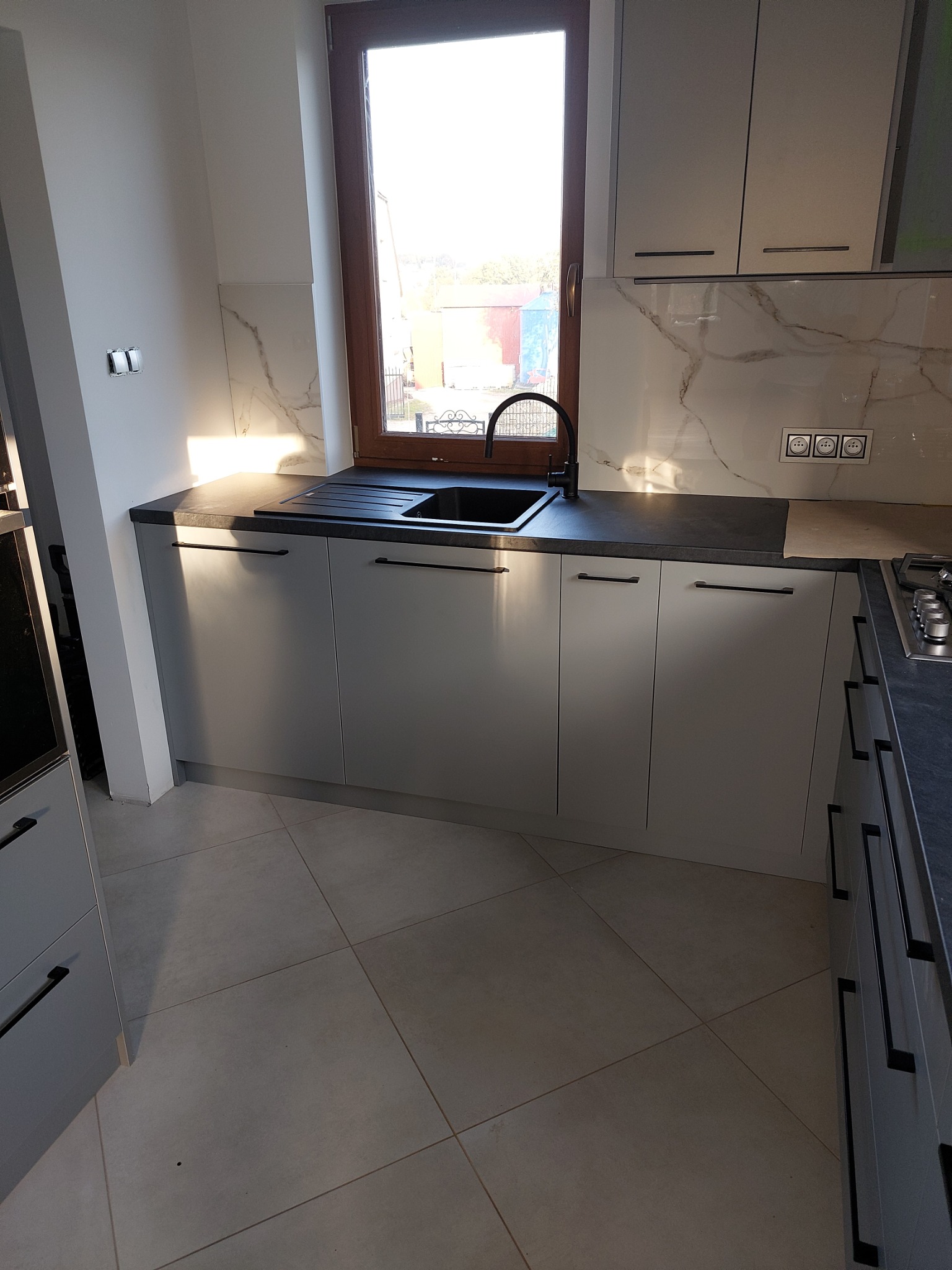 Szara zabudowa kuchenna z czarnym blatem i armaturą, marmurowy panel nad blatem. Ujęcie z perspektywy podłogi, widoczne okno. Nowoczesna kuchnia.