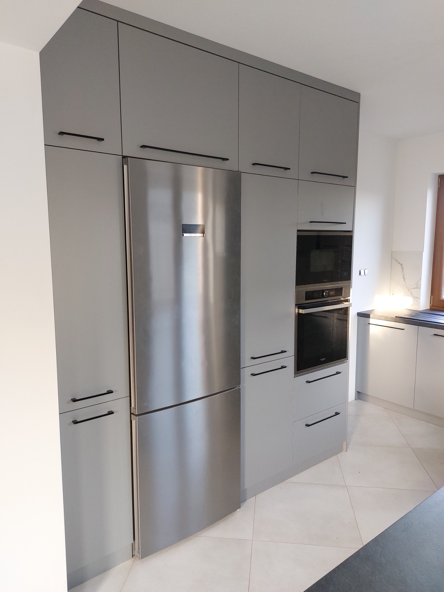 Szara zabudowa kuchenna na wymiar z lodówką i piekarnikiem w nowoczesnym stylu. Czarne, podłużne uchwyty dodają elegancji. Jasne płytki podłogowe.