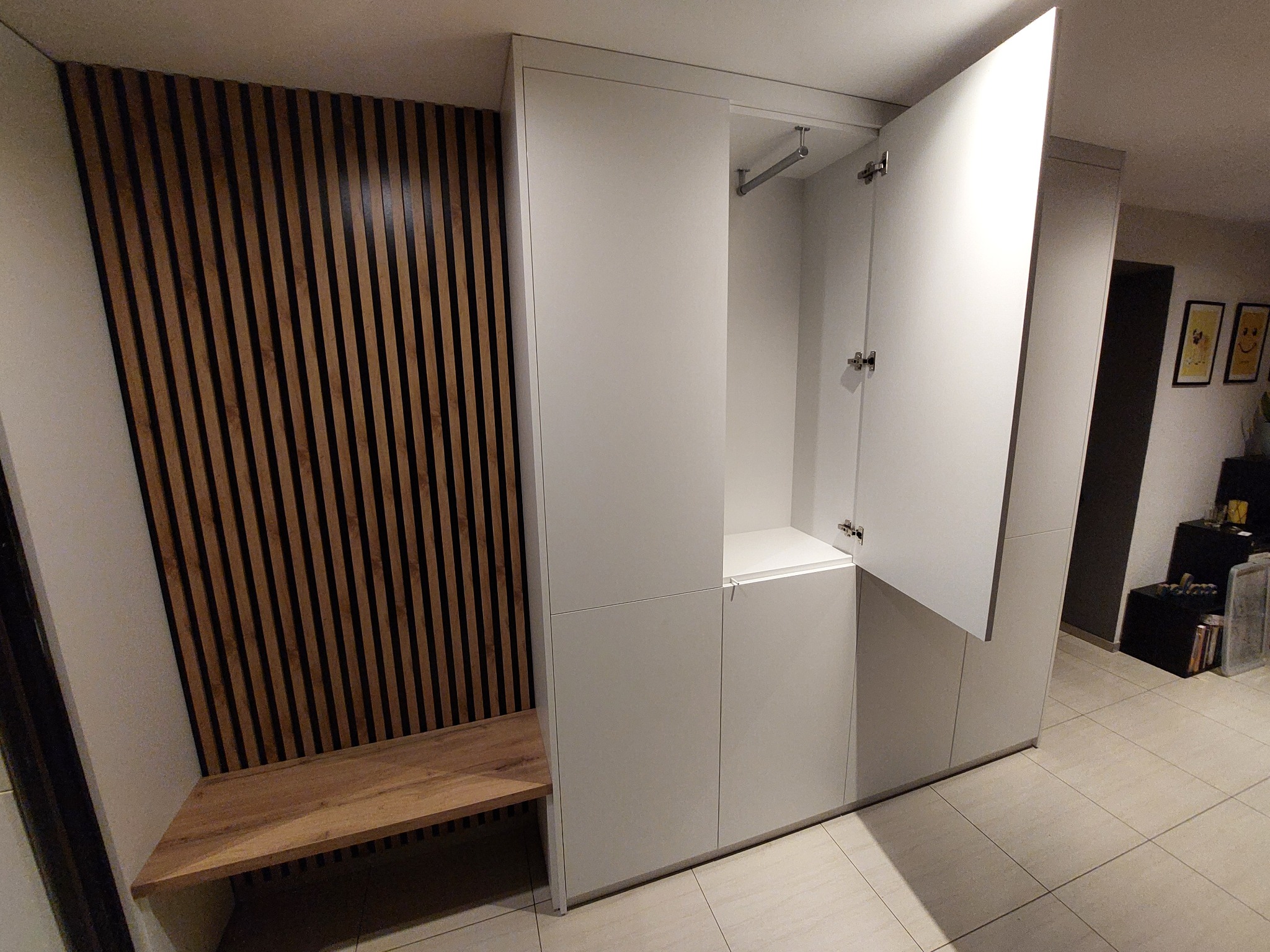 Biała szafa wnękowa z siedziskiem z drewnianych listew, otwarte drzwi ukazują drążek na ubrania. Minimalistyczny design, jasne płytki podłogowe, nowoczesne wykończenie.