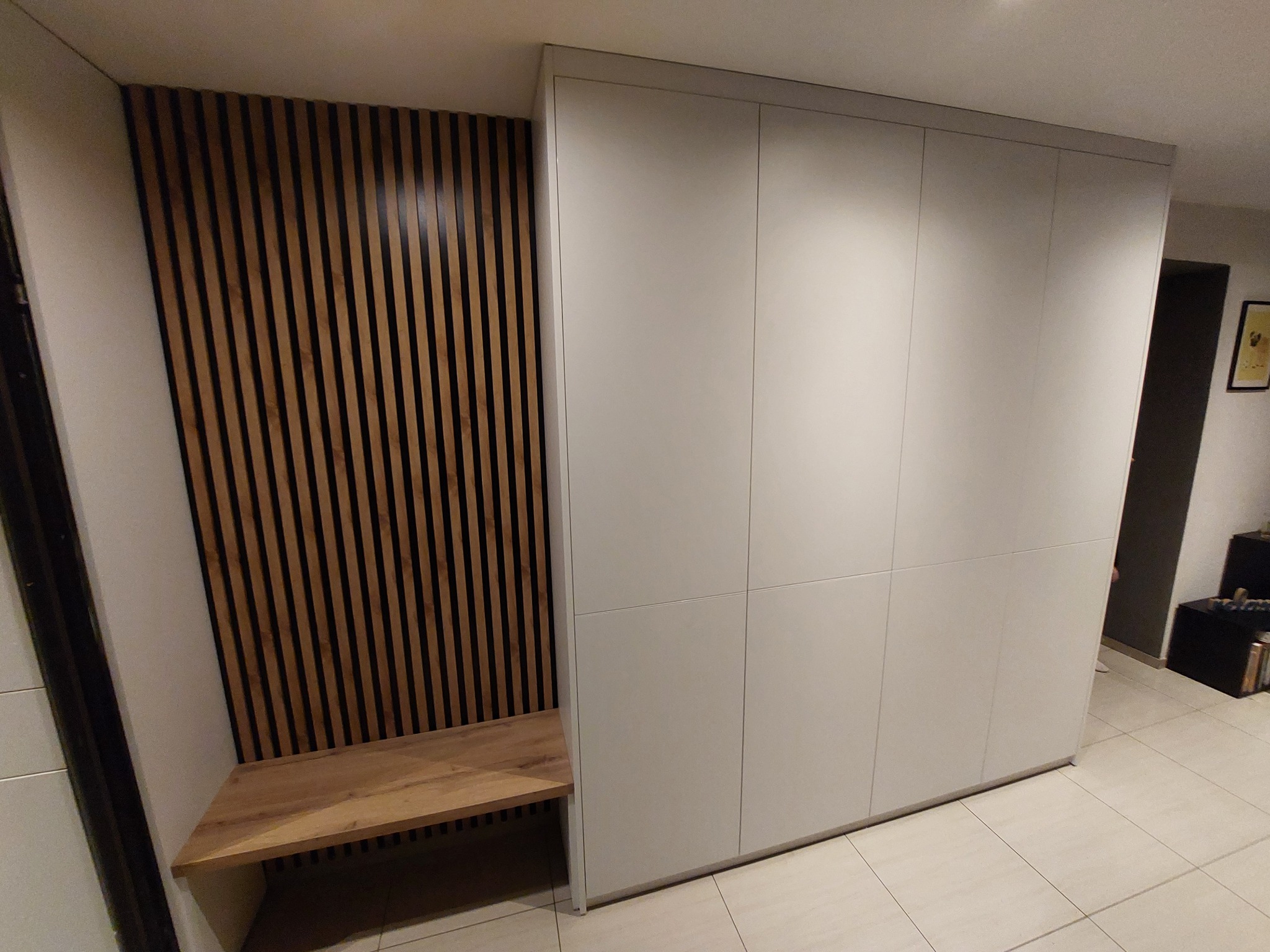 Biała szafa wnękowa z siedziskiem i dekoracyjną ścianką z drewnianych listewek w przedpokoju, widok z perspektywy podłogi, jasne płytki podłogowe.