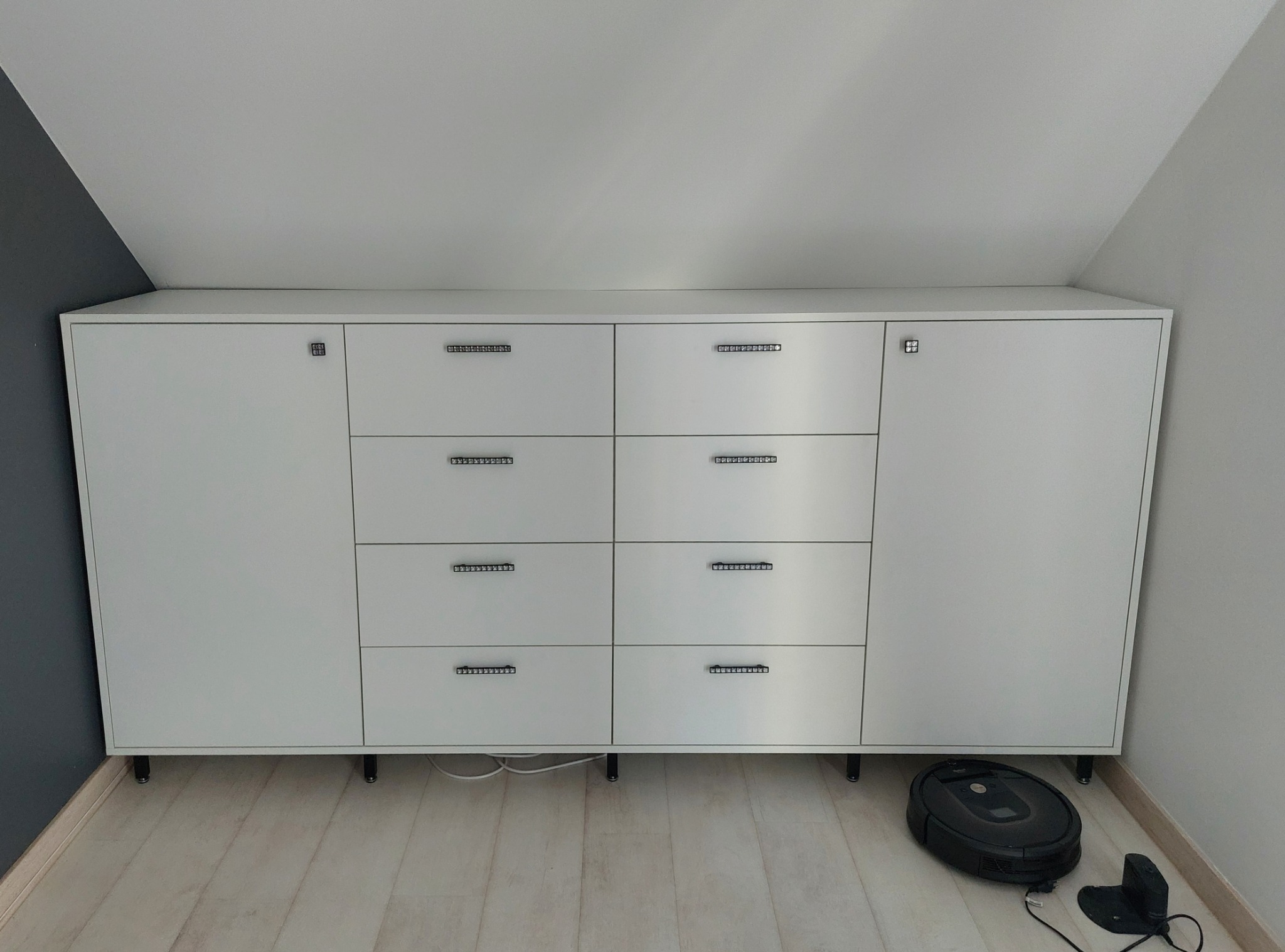 Biała szafa z szufladami i drzwiczkami, z metalowymi uchwytami, stojąca na nóżkach, w pokoju na poddaszu. Obok szafy robot sprzątający.