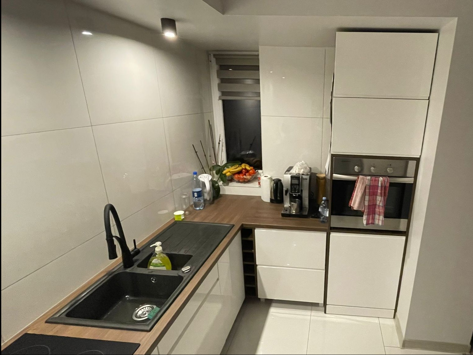 Nowoczesna kuchnia z białymi, błyszczącymi frontami, czarnym zlewem i drewnianym blatem. Widoczny piekarnik w zabudowie i okno z roletą.