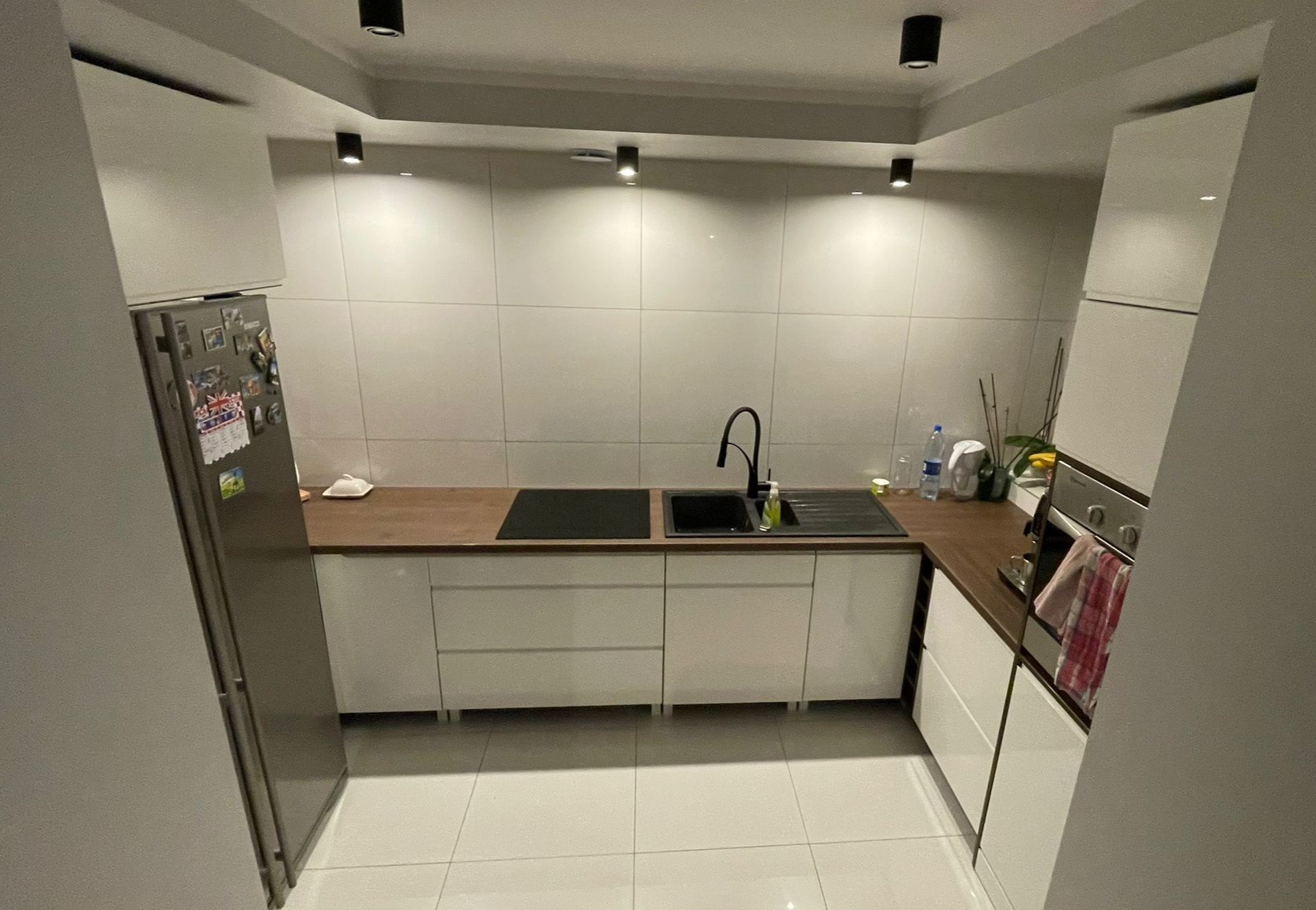 Nowoczesna, jasna kuchnia z białymi frontami, drewnianym blatem i czarną armaturą. Na lodówce magnesy. Uwagę zwraca oświetlenie sufitowe.