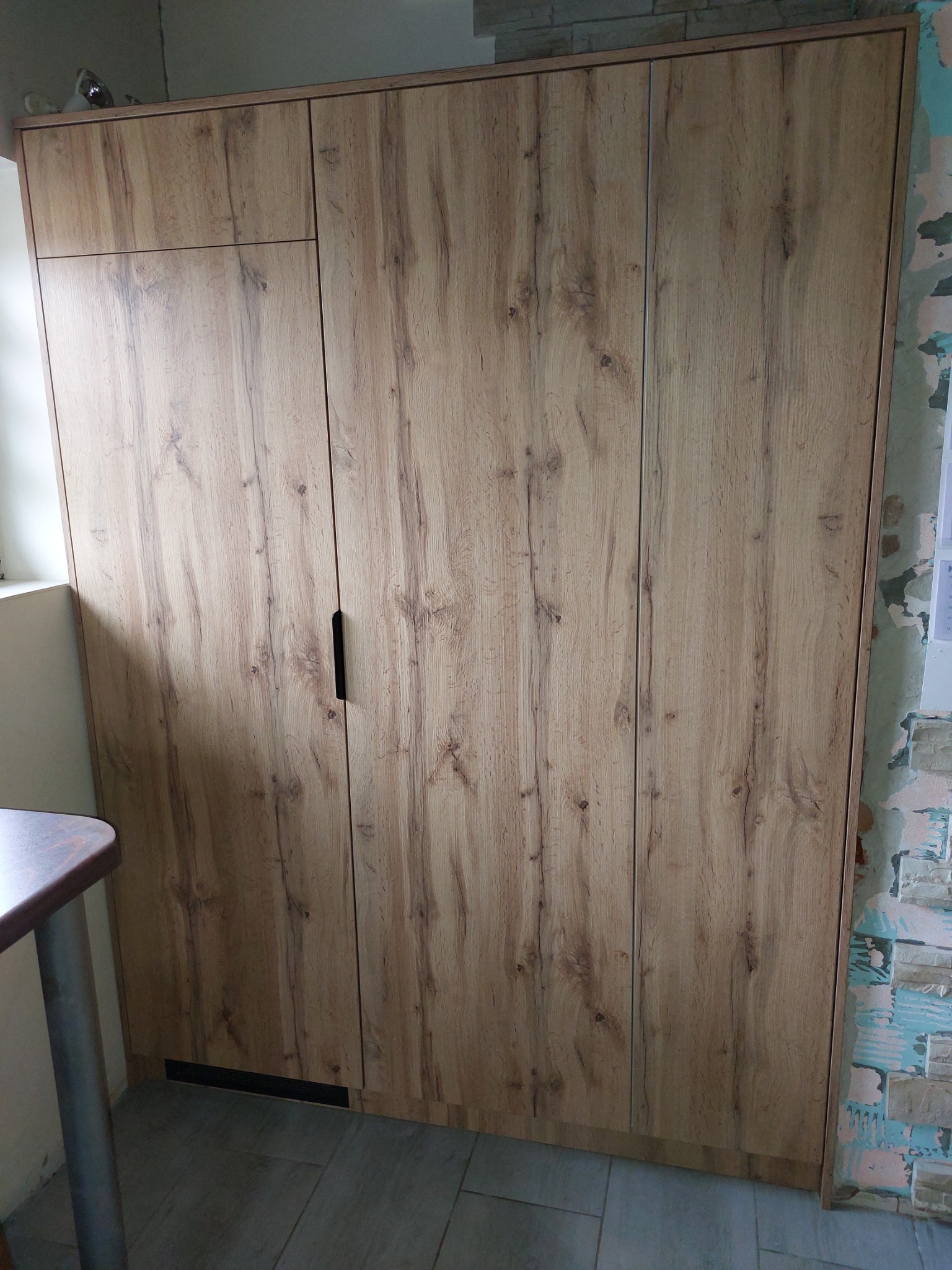 Drewniana szafa z ciemnym uchwytem, dopasowana do wnętrza. Widoczna faktura drewna i fragment ściany w tle. Szafa zamyka przestrzeń od podłogi do sufitu.