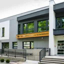 Elewacja budynku Centrum Medycznego Słowiańska z widocznym wejściem i tablicą informacyjną. Nowoczesna architektura, jasne kolory, zadbany teren.