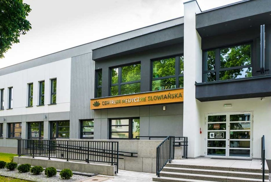 Elewacja budynku Centrum Medycznego Słowiańska z widocznym wejściem i tablicą informacyjną. Nowoczesna architektura, jasne kolory, zadbany teren.