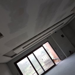 Regmal Romuald Muzika - Sufit z płyt g-k z ozdobnymi elementami w Sycowie, po szpachlowaniu, przygotowany do malowania. Widok na okno i surowy stan wykończenia.