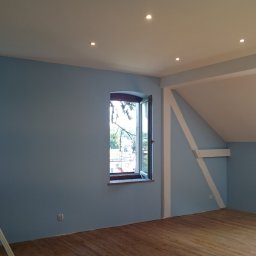 Regmal Romuald Muzika - Jasne, błękitne ściany w pokoju na poddaszu z drewnianą podłogą i oknami o różnych kształtach. Widoczne niedoskonałości malarskie przy ościeżnicy okna.