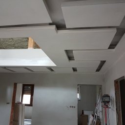 Regmal Romuald Muzika - Nowoczesny sufit podwieszany z płyt kartonowo-gipsowych w jasnym pomieszczeniu, widoczne narzędzia pracy i przygotowane płyty. Elementy dekoracyjne tworzą geometryczny wzór.