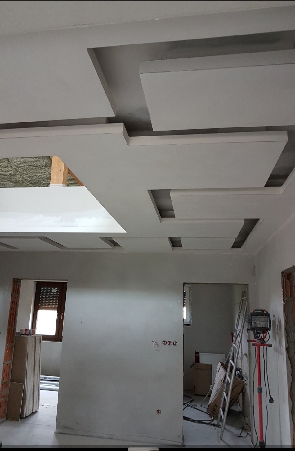 Nowoczesny sufit podwieszany z płyt karton-gips, z geometrycznymi wzorami i wnękami, oświetlenie halogenowe w tle, wykończenie wnętrza w trakcie realizacji.