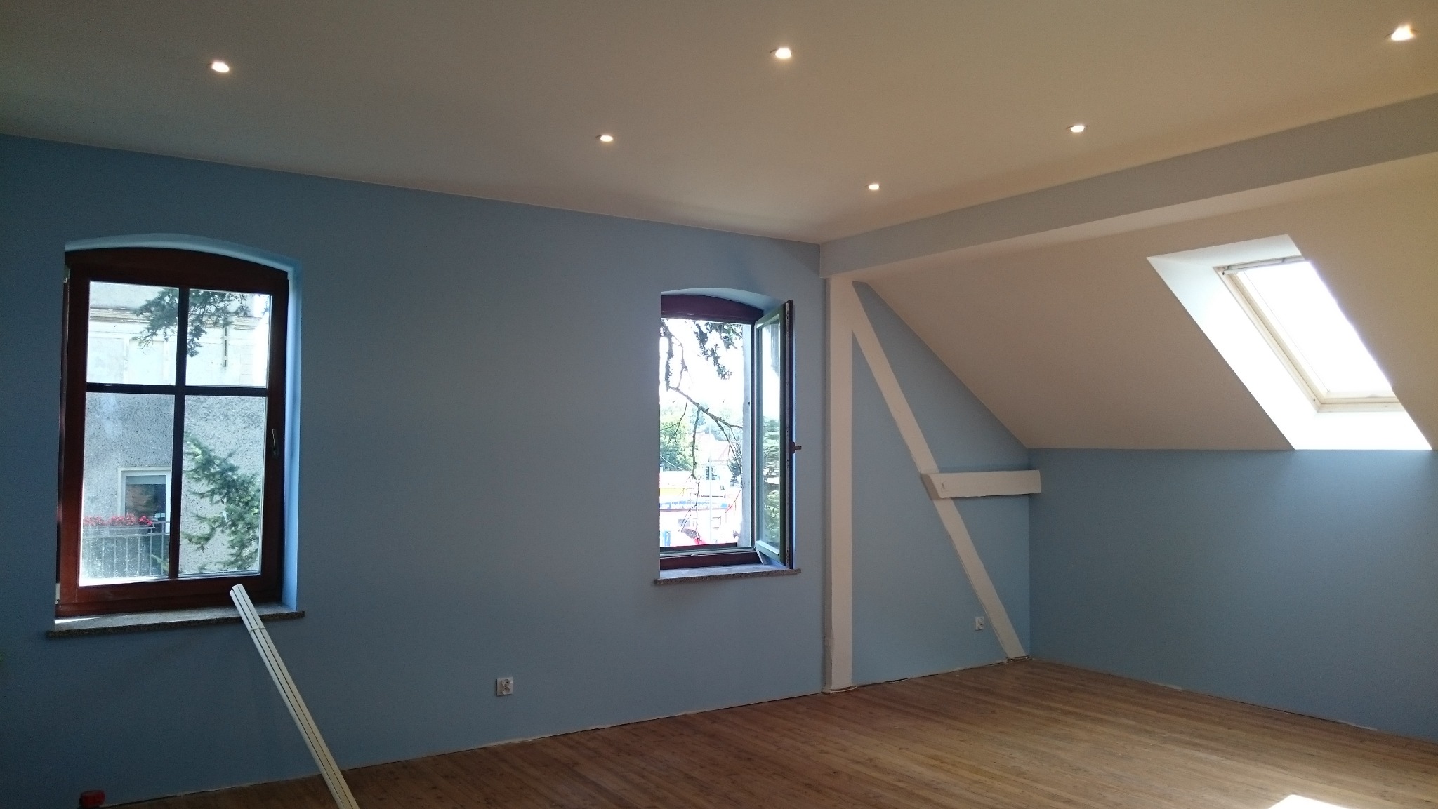 Jasne, błękitne ściany w pokoju na poddaszu z drewnianą podłogą i oknami o różnych kształtach. Widoczne niedoskonałości malarskie przy ościeżnicy okna.