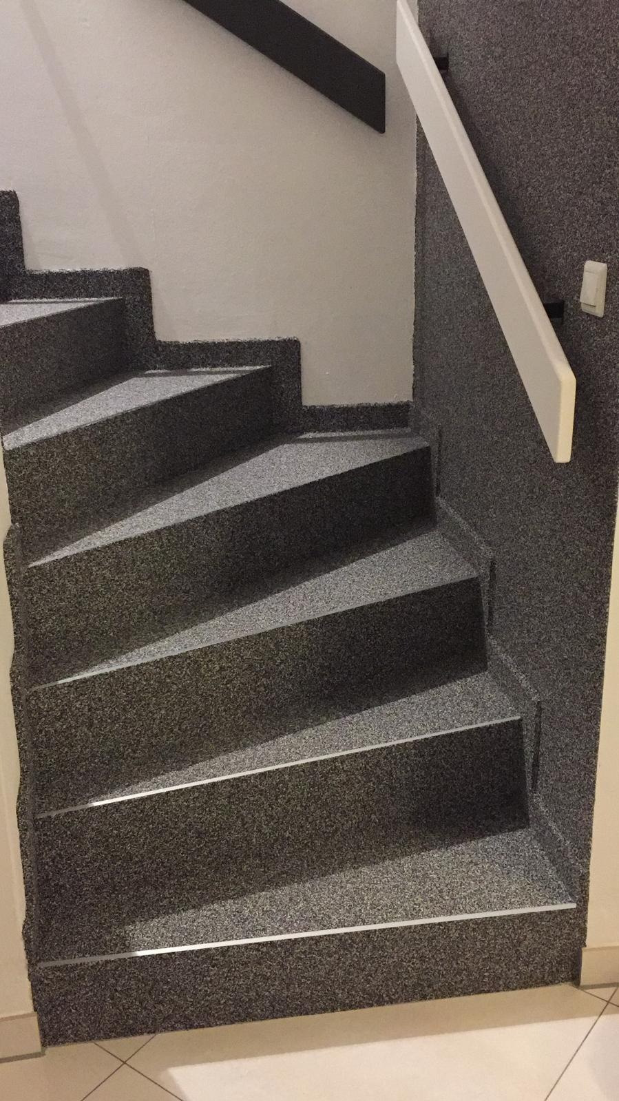 Schody wykończone szarą wykładziną dywanową z białymi poręczami i aluminiowymi listwami na krawędziach stopni, widok z dołu, jasne płytki na podłodze.