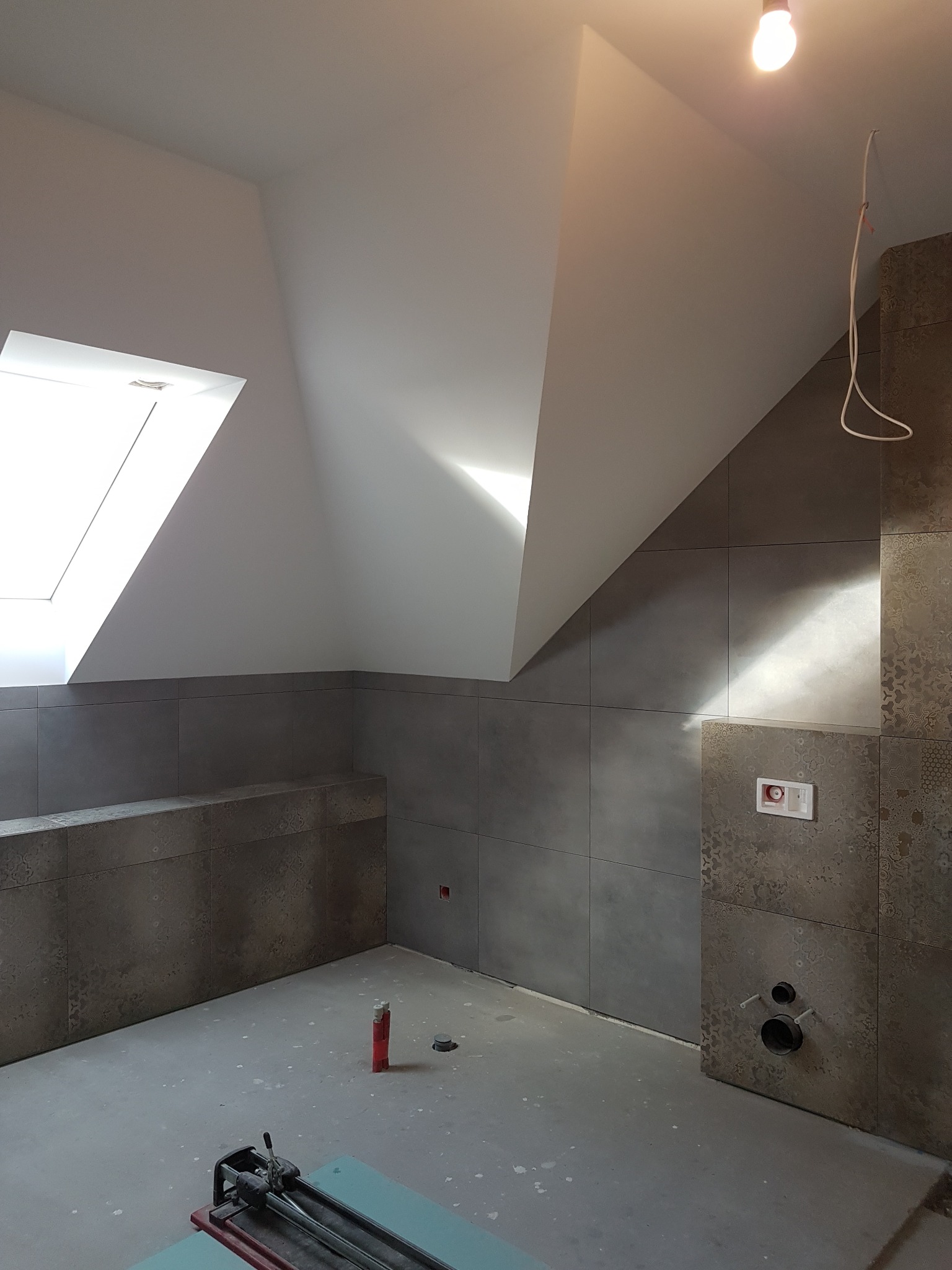 Wykończona łazienka na poddaszu z oknem dachowym. Szare płytki na ścianach, widoczne przyłącza wod-kan, narzędzie do cięcia płytek na podłodze.