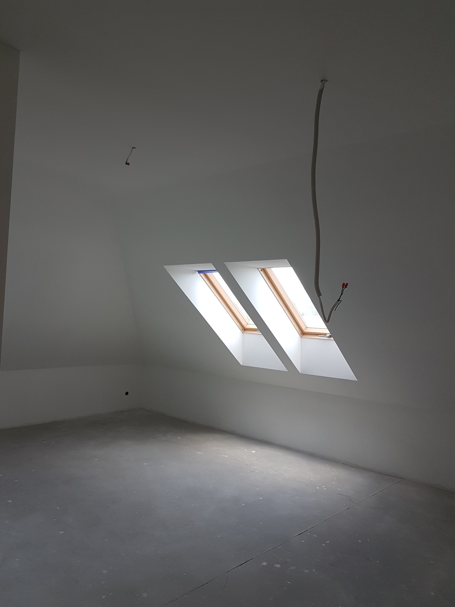 Jasne poddasze po remoncie z dwoma oknami dachowymi i wystającym przewodem elektrycznym. Ściany i podłoga w jednolitym, jasnym kolorze.