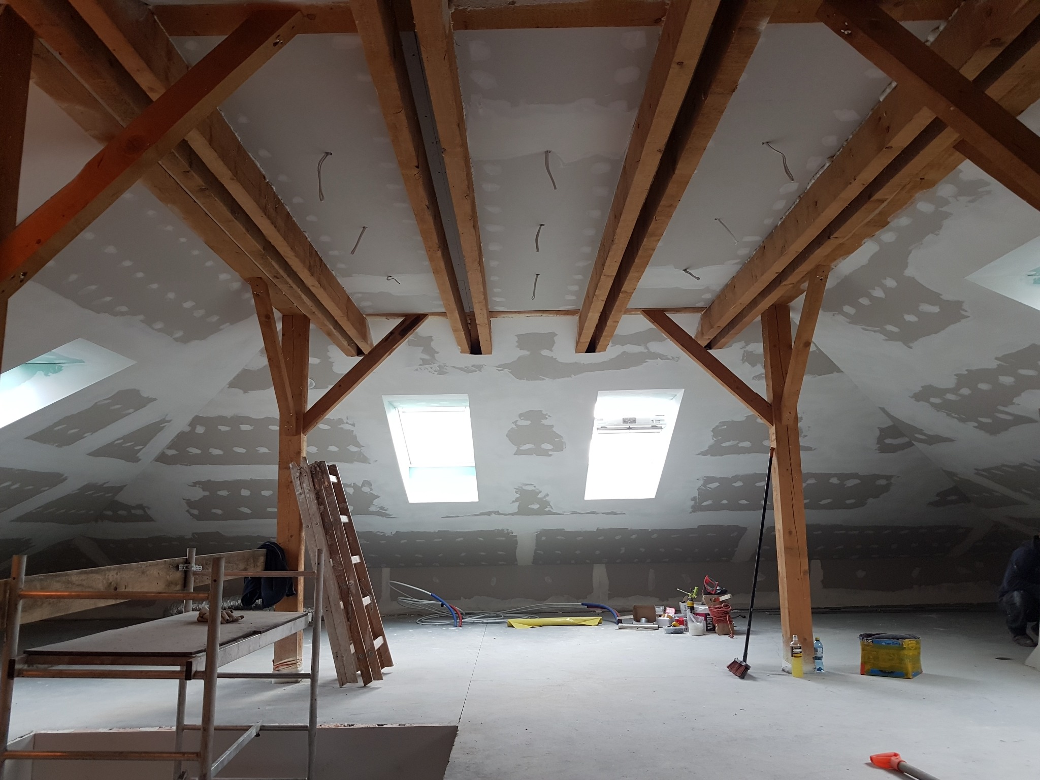 Poddasze w trakcie prac wykończeniowych: widoczne drewniane belki, okna dachowe, szpachlowane ściany i sufit z płyt gipsowo-kartonowych. Rozstawiony ruszt i drabina.