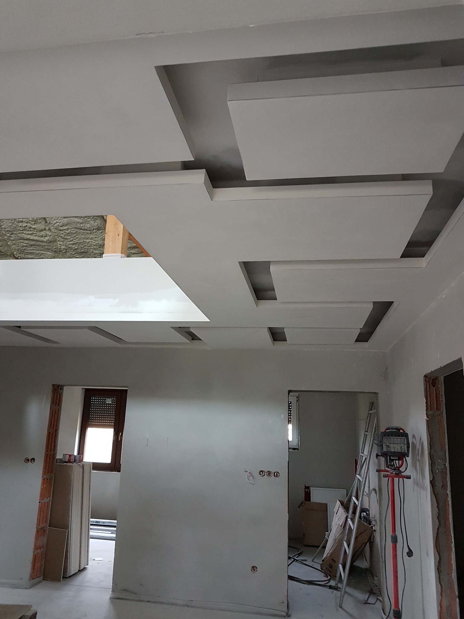 Nowoczesny sufit podwieszany z płyt kartonowo-gipsowych w jasnym pomieszczeniu, widoczne narzędzia pracy i przygotowane płyty. Elementy dekoracyjne tworzą geometryczny wzór.