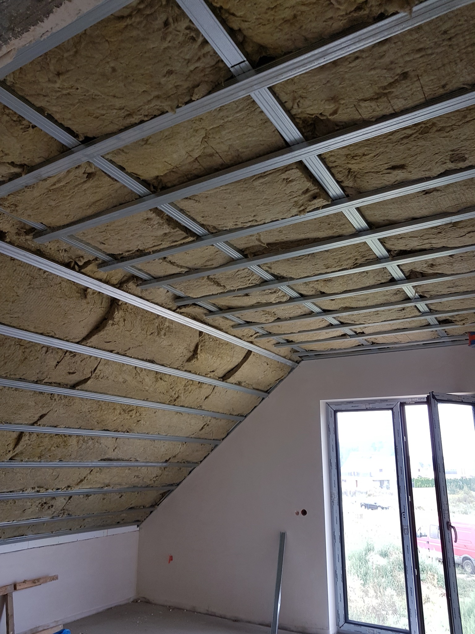 Montaż izolacji z wełny mineralnej w metalowym stelażu sufitu poddasza. Widoczne okno i fragment ściany. Etap prac wykończeniowych w nowym budynku.