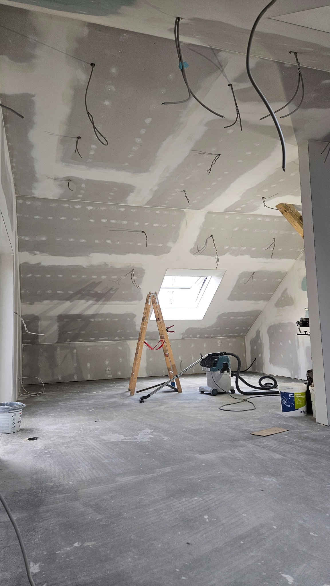 Poddasze po szpachlowaniu płyt gipsowych, widoczne kable, drabina i odkurzacz przemysłowy. Sufit ze świetlikiem. Ściany przygotowane do malowania.
