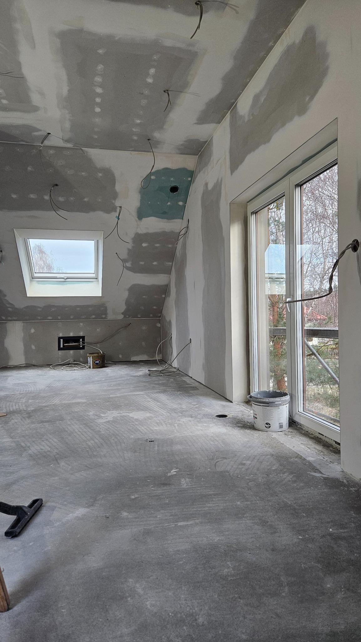 Pokój na poddaszu w trakcie szpachlowania: ściany i sufit pokryte szpachlą, widoczne kable, okno dachowe i okno balkonowe. Na podłodze widoczne ślady szpachli.