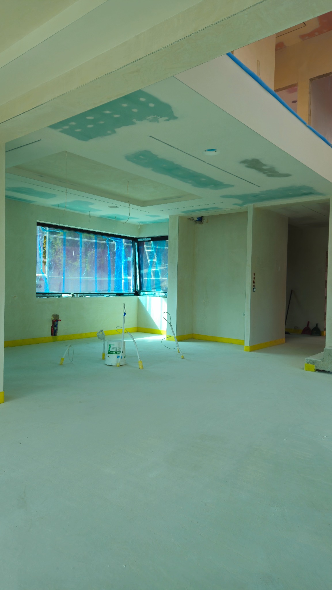 Wnętrze w trakcie prac wykończeniowych: sufit z płyt karton-gips, zabezpieczona folią okienna rama, podłoga przygotowana do dalszych prac, widoczne narzędzia malarskie.