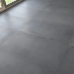 Ułożona, ciemnoszara, matowa podłoga z płytek w pomieszczeniu z oknem balkonowym. Widoczne fugi i fragment białej ściany w rogu.