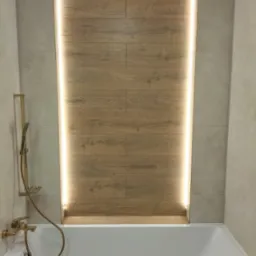 Nowoczesna łazienka z wanną, beżowymi płytkami i drewnopodobną ścianą z podświetleniem LED. Złota armatura dodaje elegancji.