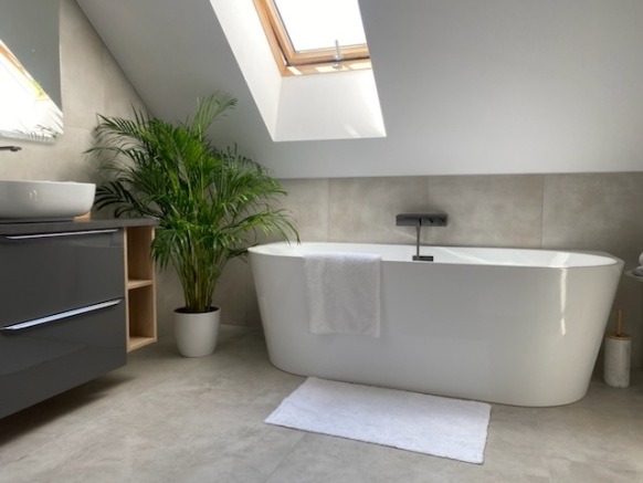 Jasna łazienka na poddaszu z wanną, umywalką i rośliną doniczkową. Minimalistyczny design, szare płytki, okno dachowe. Nowoczesne wykończenie.
