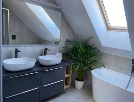 Jasna łazienka na poddaszu z dwoma umywalkami na szafce, wanną i oknem dachowym, doświetlona naturalnym światłem. Wnętrze uzupełnia zielona roślina doniczkowa.