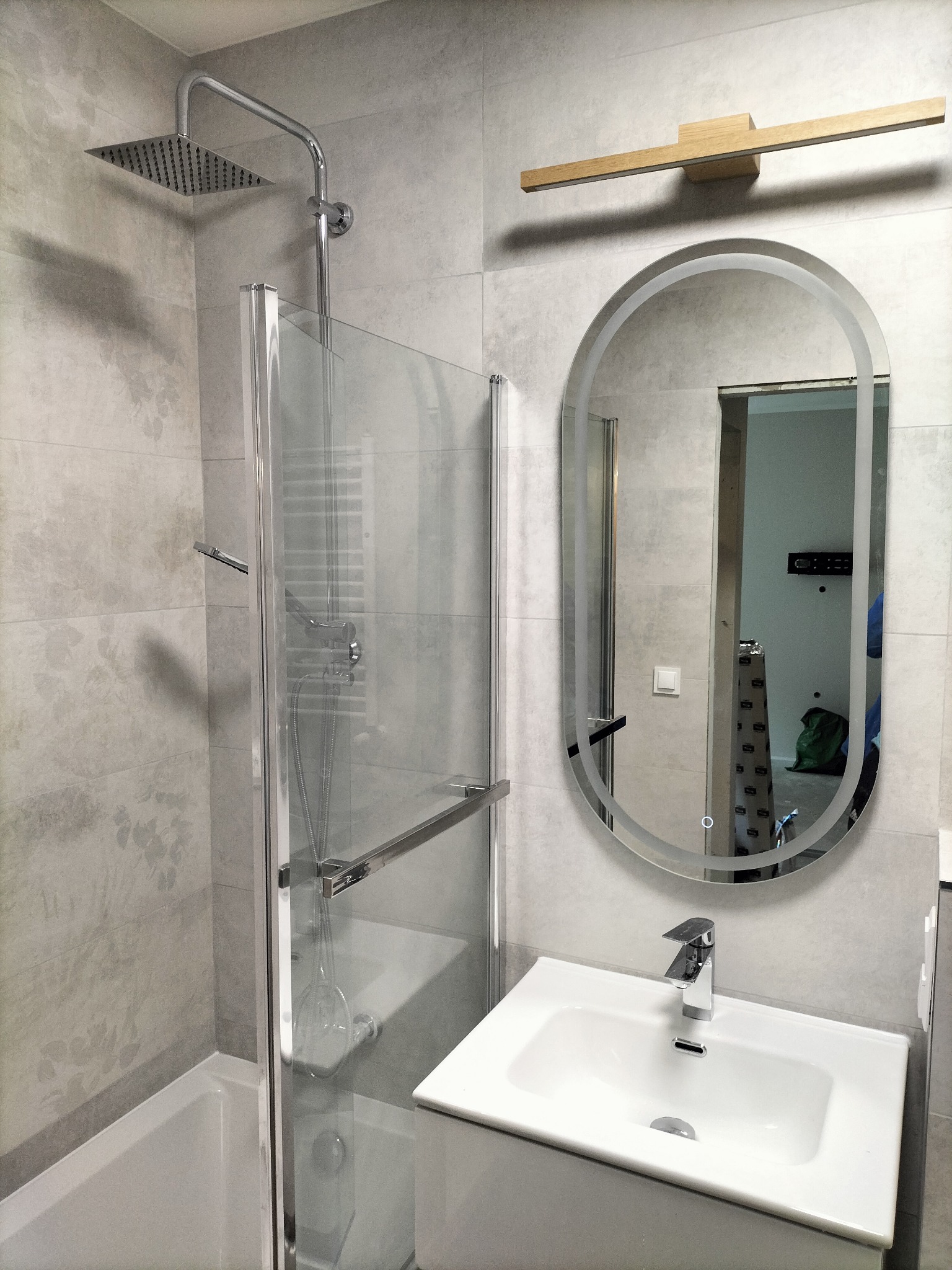 Nowoczesna łazienka z kabiną prysznicową typu walk-in, owalnym lustrem z podświetleniem LED i drewnianym oświetleniem nad nim. Szare płytki z delikatnym wzorem.