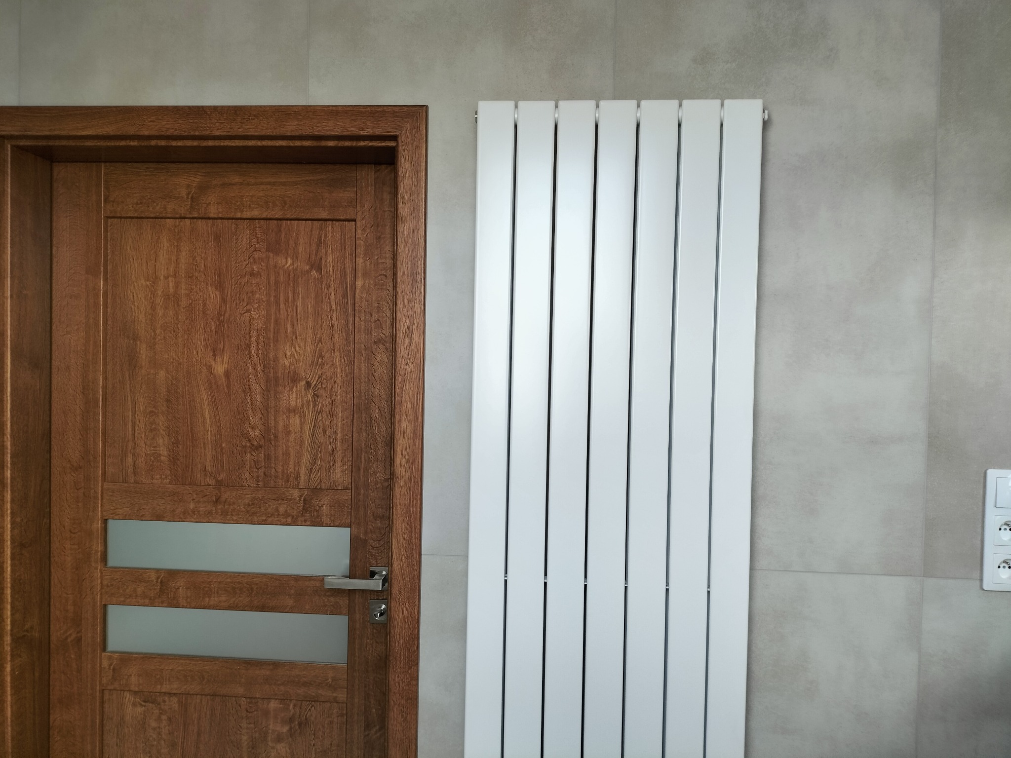 Nowoczesna łazienka: drewniane drzwi z przeszkleniami i biały, pionowy grzejnik na tle szarej, kwadratowej glazury ściennej, z widocznym gniazdkiem elektrycznym.