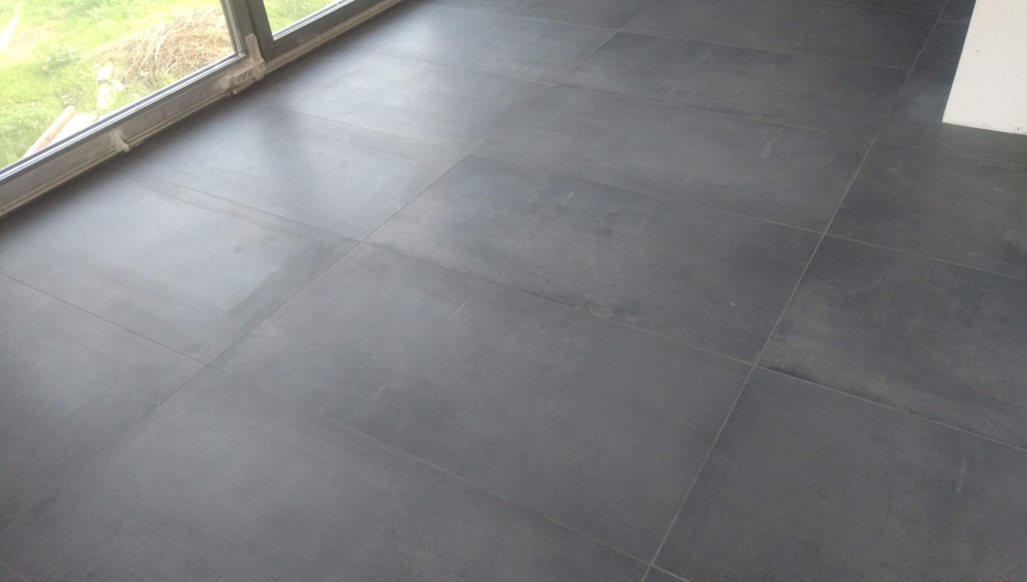 Ułożona, ciemnoszara, matowa podłoga z płytek w pomieszczeniu z oknem balkonowym. Widoczne fugi i fragment białej ściany w rogu.