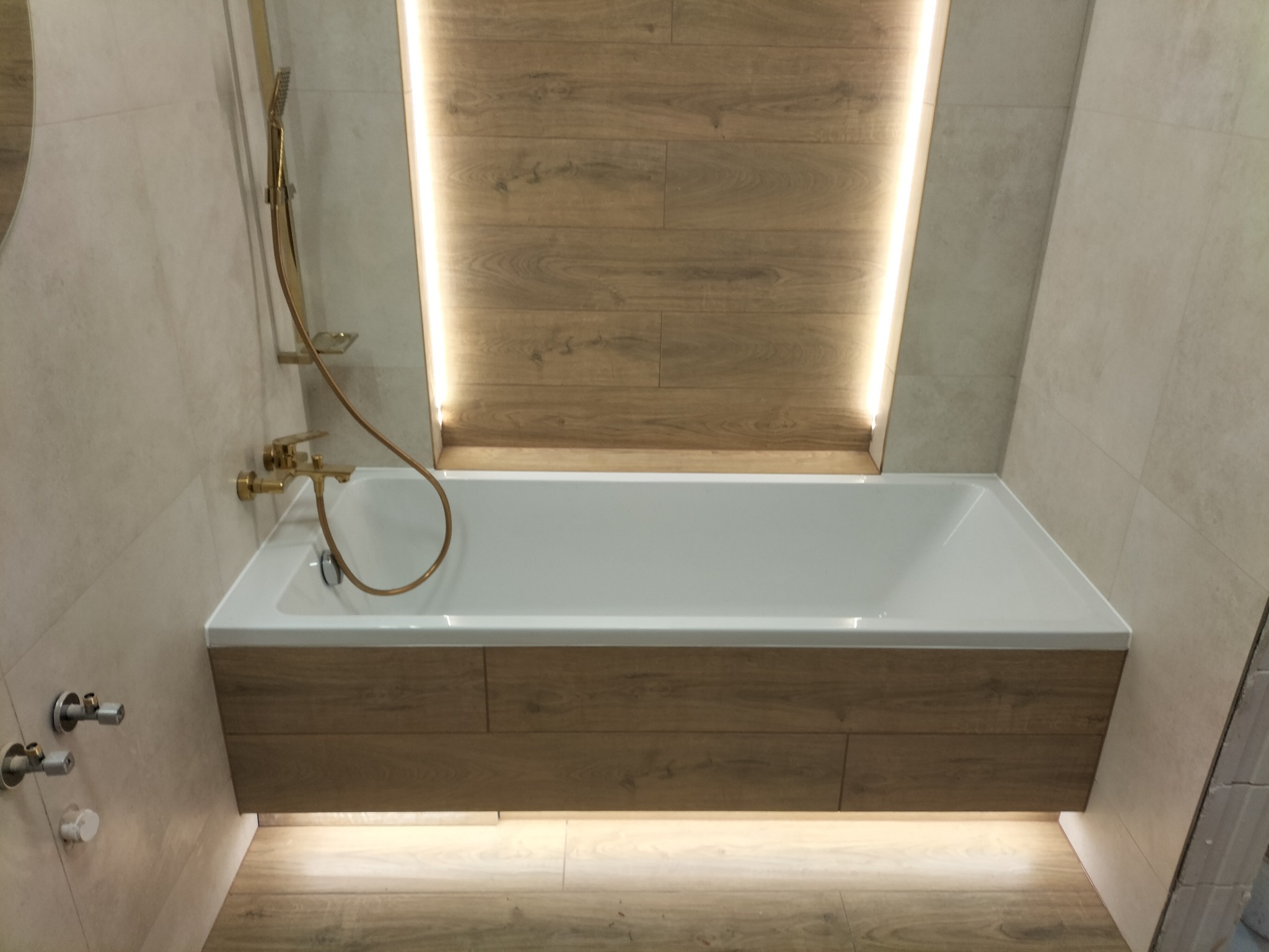 Nowoczesna łazienka: biała wanna obudowana drewnem, złota armatura, podświetlenie LED i płytki w odcieniach beżu tworzą elegancką przestrzeń kąpielową.