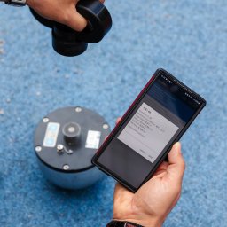 EUROPEJSKIE CENTRUM JAKOŚCI I PROMOCJI SPÓŁKA Z OGRANICZONĄ ODPOWIEDZIALNOŚCIĄ - Pomiar HIC (Head Injury Criterion) na placu zabaw. Telefon z aplikacją prezentuje wyniki testu, obok urządzenie pomiarowe. Badanie bezpieczeństwa nawierzchni.