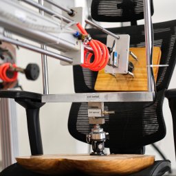 EUROPEJSKIE CENTRUM JAKOŚCI I PROMOCJI SPÓŁKA Z OGRANICZONĄ ODPOWIEDZIALNOŚCIĄ - Urządzenie testujące wytrzymałość krzesła biurowego z czujnikami i siłownikami, na tle ściany z logotypem. Testy ergonomii i wytrzymałości mebli biurowych.