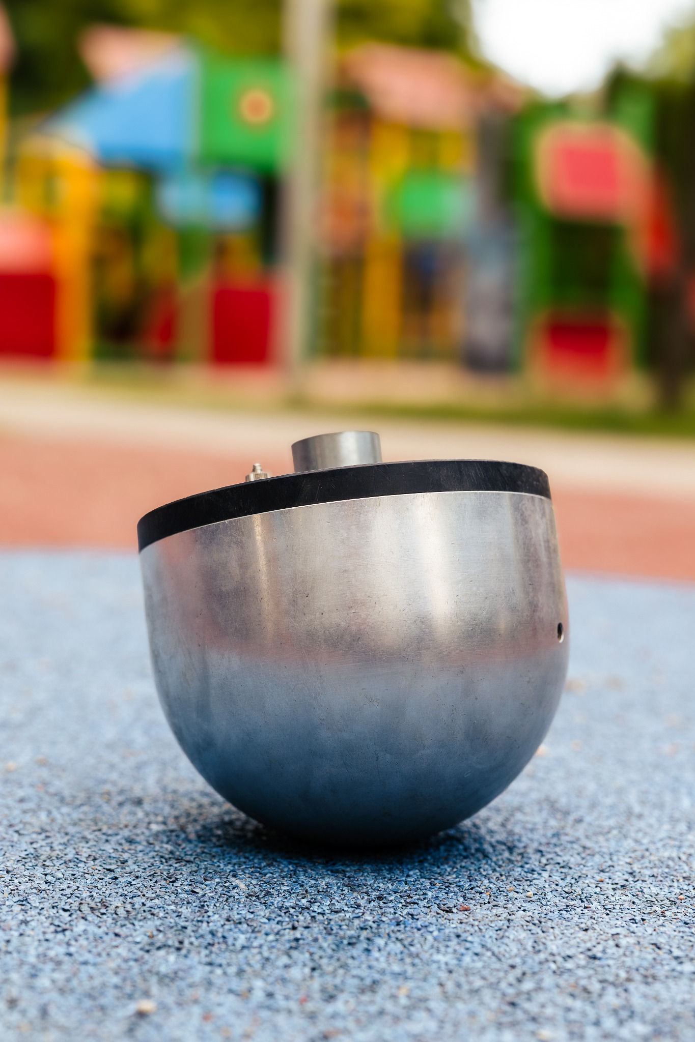 Metalowa półkula z otworem na tle placu zabaw. Urządzenie na placu zabaw dla dzieci, wykonane ze stali nierdzewnej, umieszczone na niebieskiej, gumowej nawierzchni.