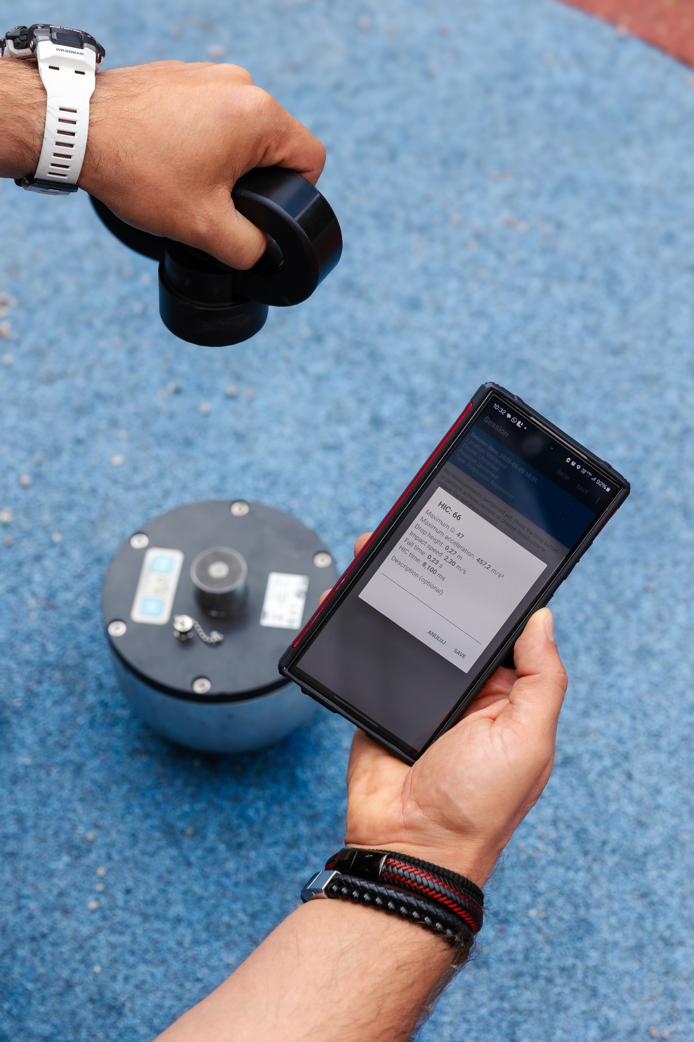 Pomiar HIC (Head Injury Criterion) na placu zabaw. Telefon z aplikacją prezentuje wyniki testu, obok urządzenie pomiarowe. Badanie bezpieczeństwa nawierzchni.