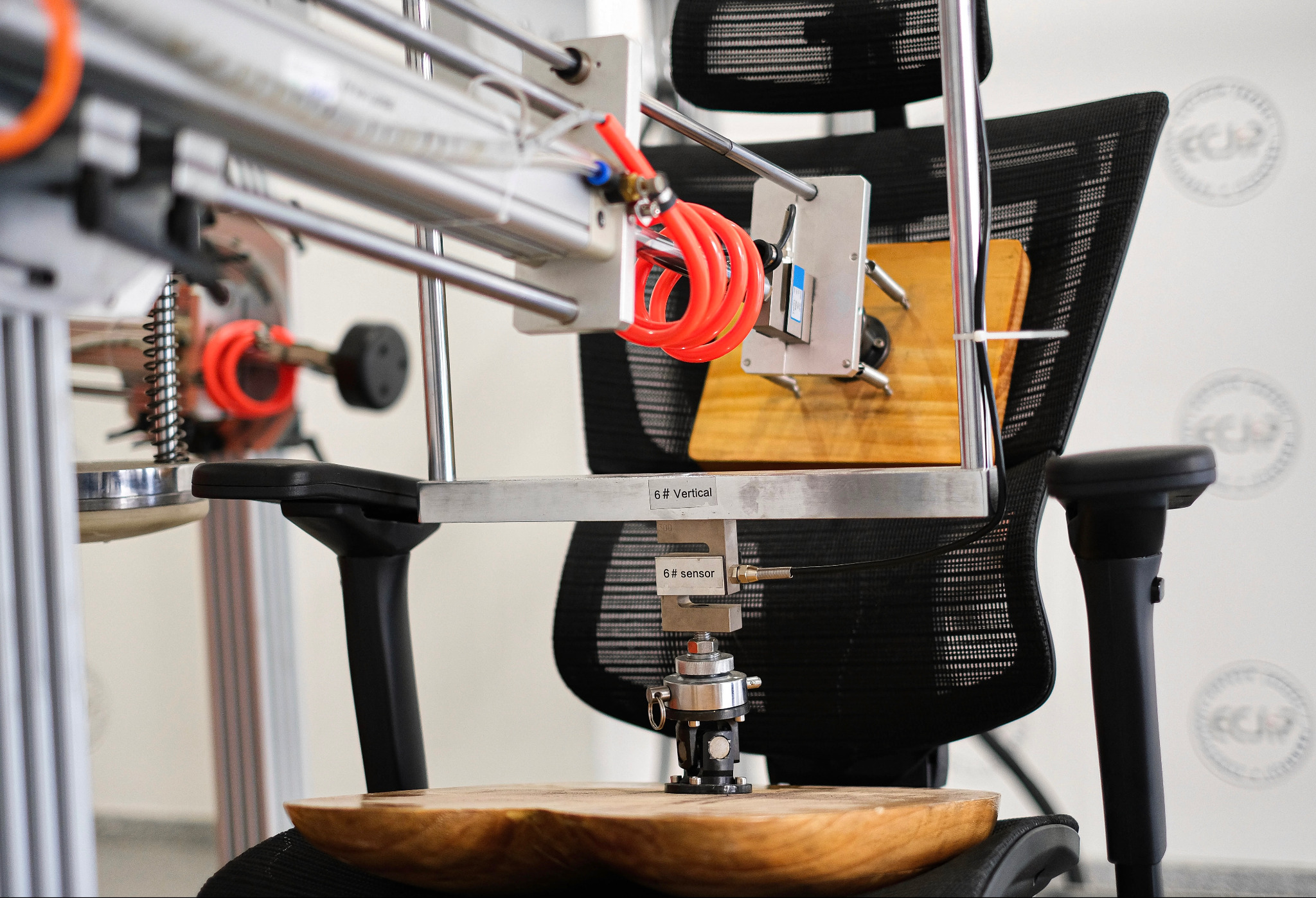 Urządzenie testujące wytrzymałość krzesła biurowego z czujnikami i siłownikami, na tle ściany z logotypem. Testy ergonomii i wytrzymałości mebli biurowych.