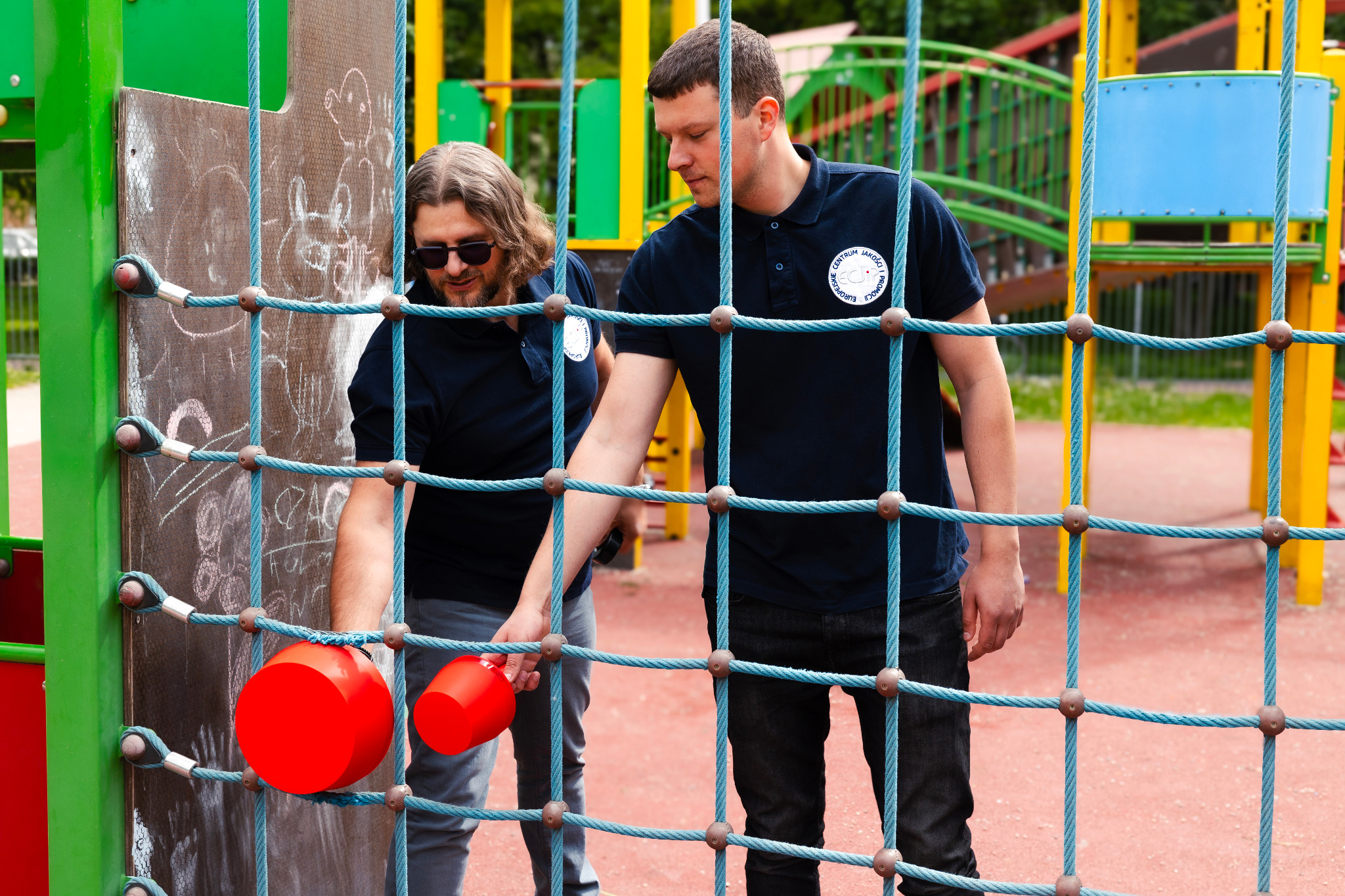 Dwóch mężczyzn w granatowych koszulkach z logo, testuje sprawność zabawki na placu zabaw, sprawdzając certyfikację bezpieczeństwa.